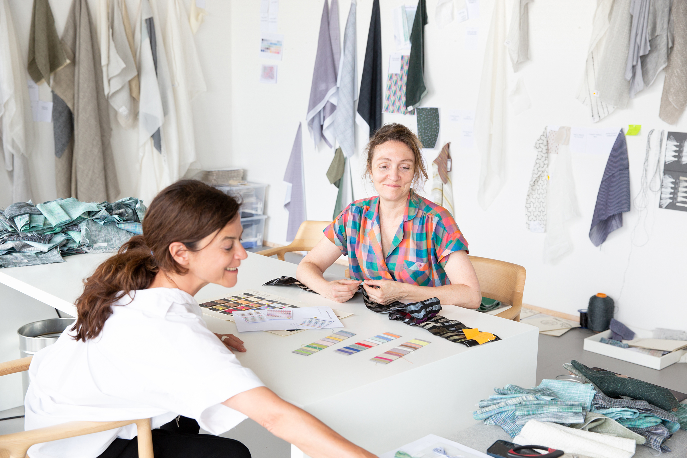 Isa Glink and Inga Sempé sitting at a table with fabric samples, colors, and patterns, with additional fabrics and designs hanging in the background.