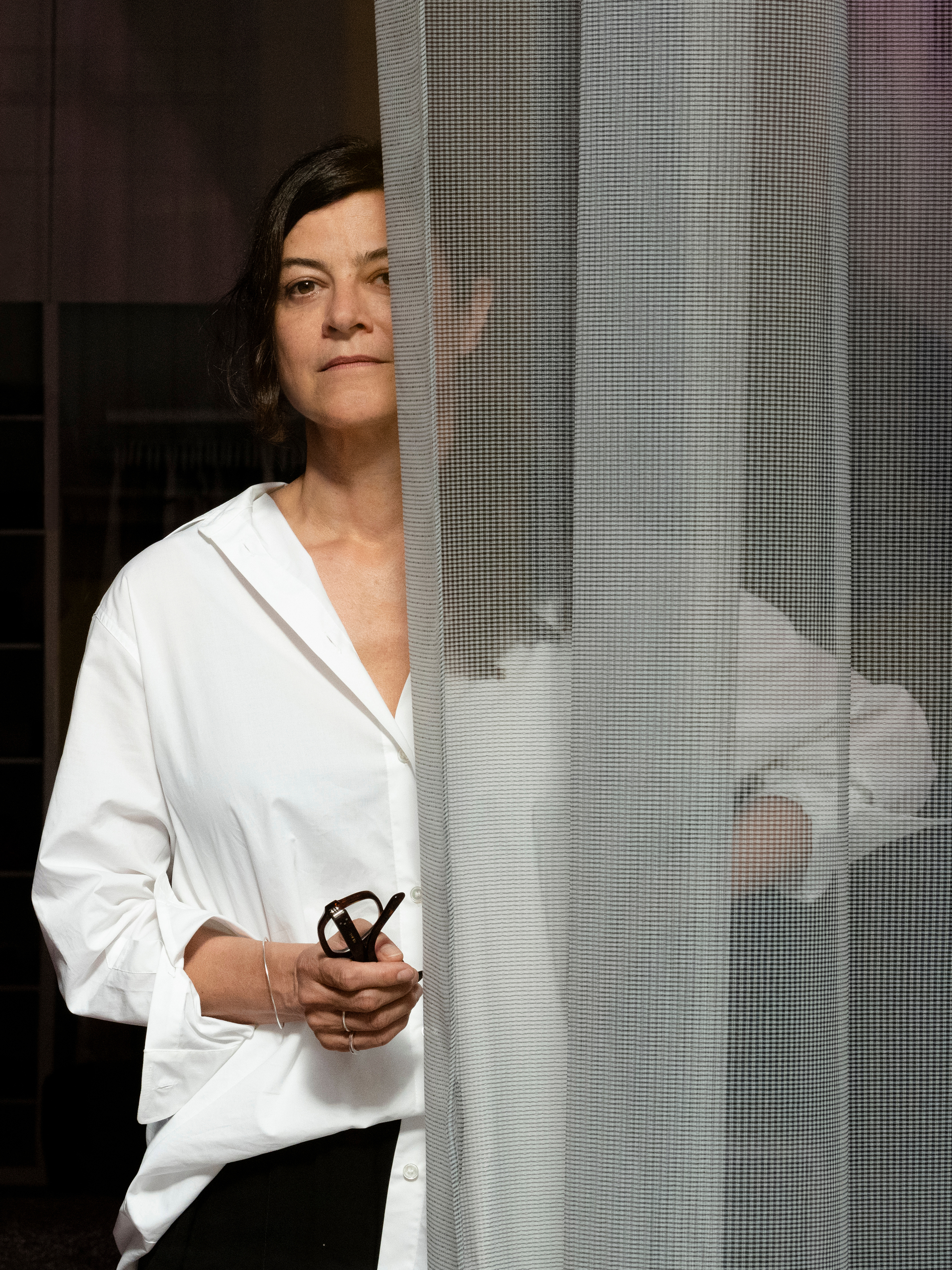 Portrait of Isa Glink, standing partially behind a semi-transparent curtain, holding glasses in her hand.