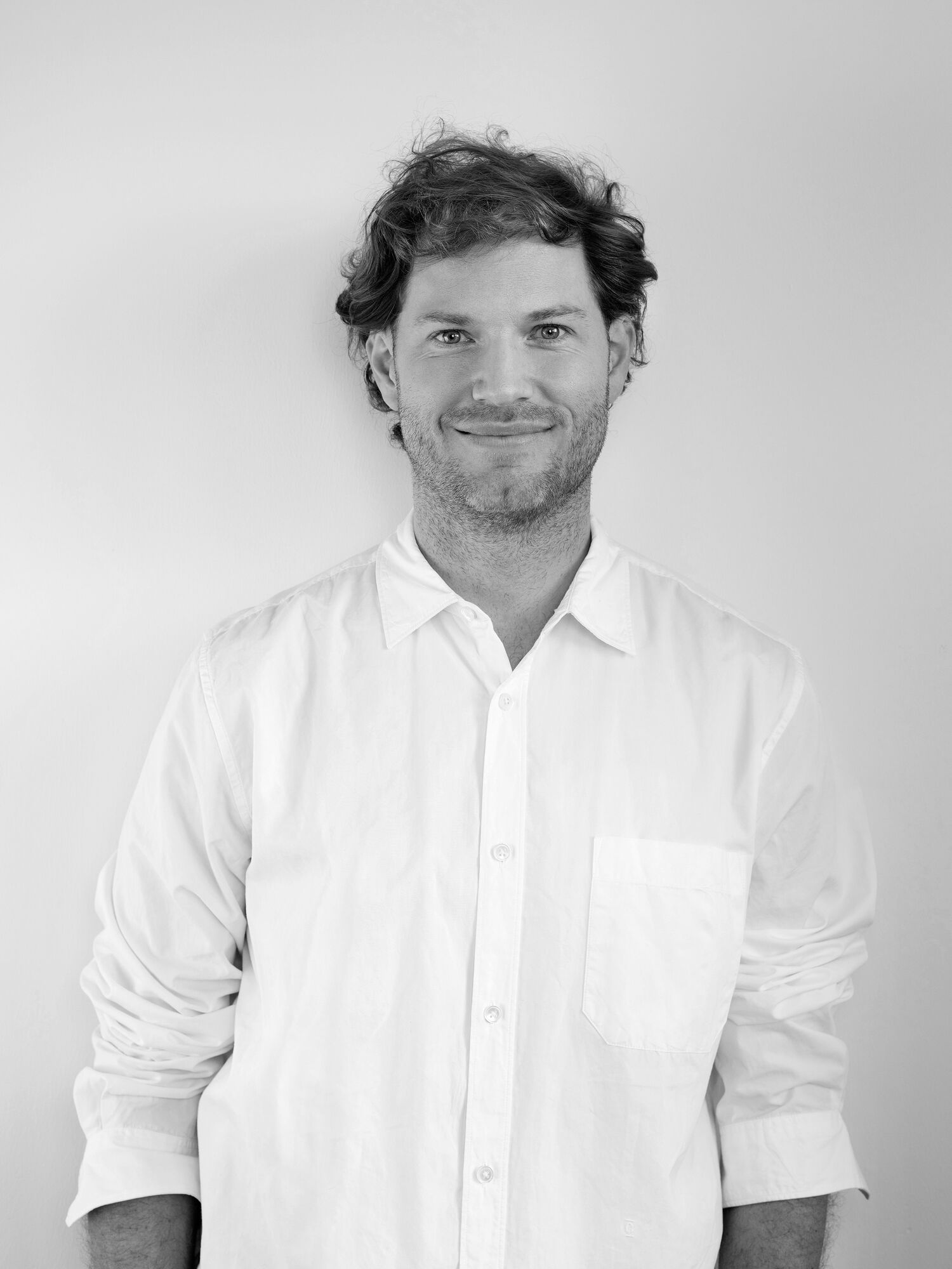 Schwarz-weiß-Portrait von Maximilian Bülk in weißem Hemd, lächelnd, mit dem Rücken an eine Wand gelehnt.