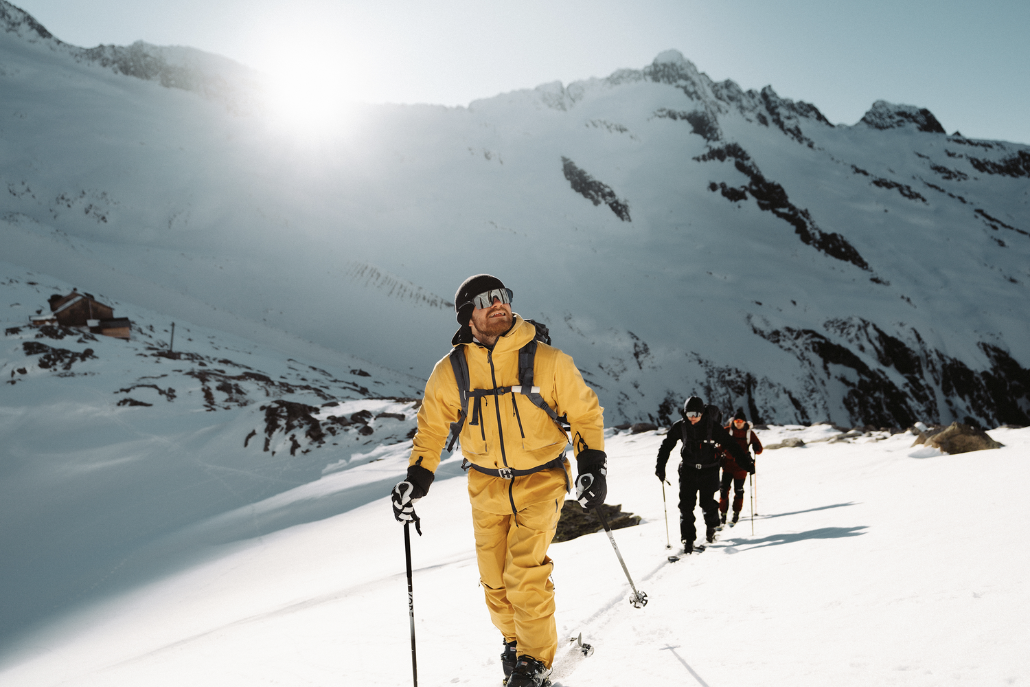Drei Personen wandern in Reihe durch eine verschneite Berglandschaft. Sie haben Wanderstöcke und Skibrillen. Die Person ganz vorne ist in Gelb gekleidet. Die beiden anderen in Schwarz.