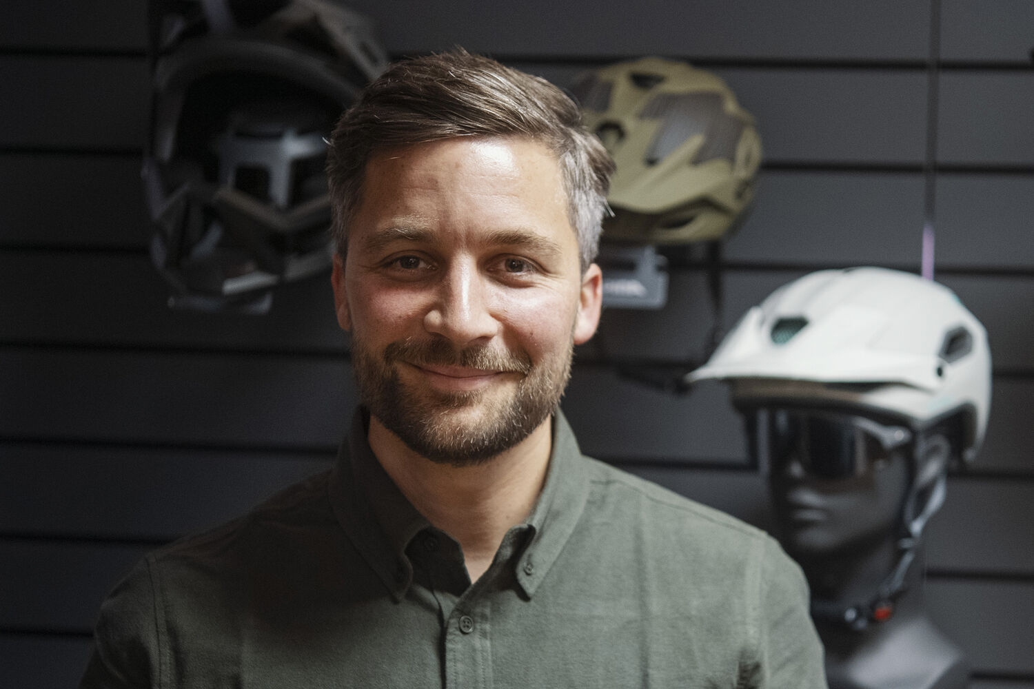 Ein Porträt von Moritz Maier. Er ist weiß, hat volles Braun-Graues Haar, einen Vollbart, dunkle Augen und er trägt ein grünes Hemd. Er lächelt leicht. Er befindet sich vor einer schwarzen Wand mit Outdoor-Helmen und -Brillen.