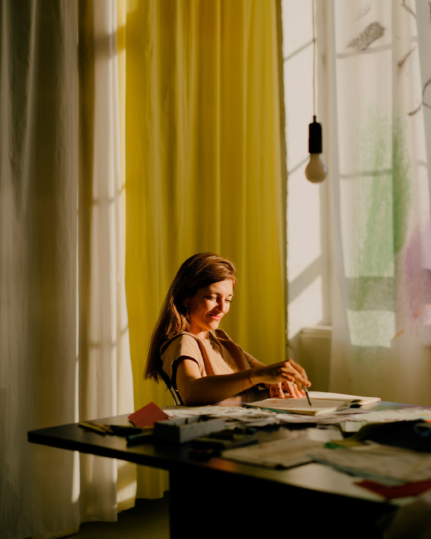 Smiling woman sits at a table and draws. Around her are hung different curtains in front of the windows. Photography for the campaign.