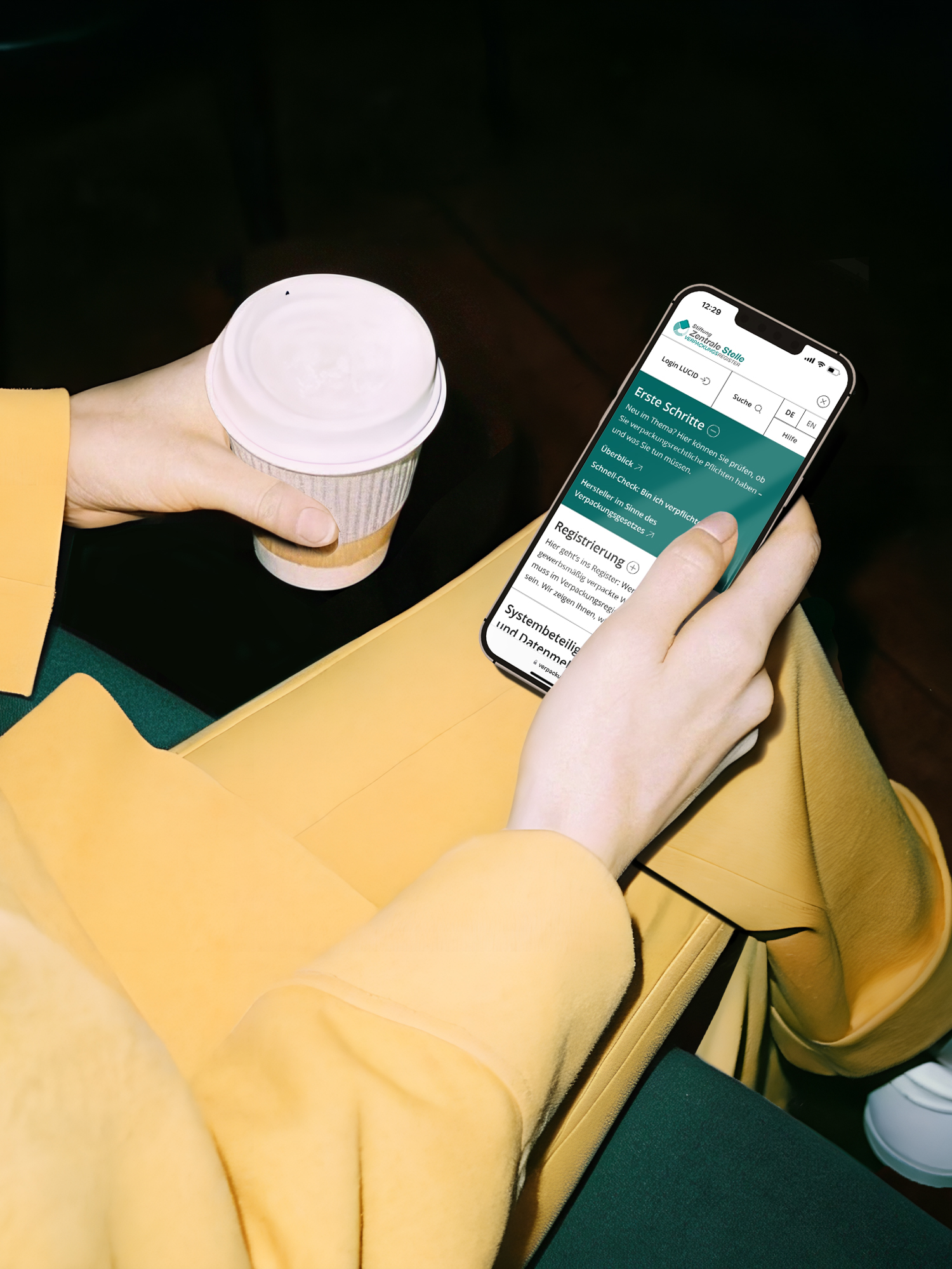 Eine Frau in einem gelben Anzug hält ein Smartphone mit geöffnetem Menü der ZSVR Website in der einen Hand und einen Coffee-To-Go-Becher in der anderen.